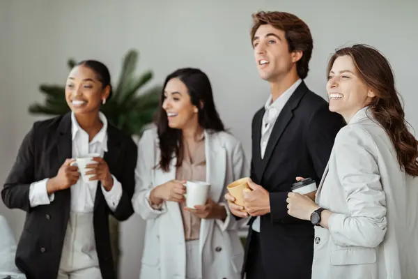 Group of happy international colleagues having break, drinking coffee, holding cups and talking, looking together aside and laughing in office interior