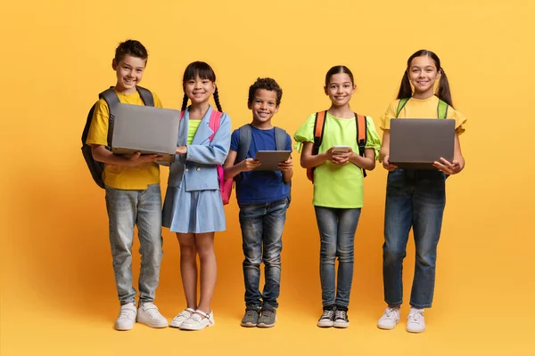 かわいい多民族の子供たちは 黄色の背景に隔離されたラップトップコンピュータPc デジタルタブレット 携帯電話を保持するバックパックを持つ幸せな少年や少女たち 素晴らしい教育ウェブサイト アプリケーションを使用して子供たち — ストック写真