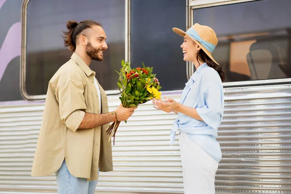 Cheerful young caucasian guy meets surprised woman, gives bouquet of flowers on train station. Hello, love, relationship, travel and lifestyle, dating and romance, vacation