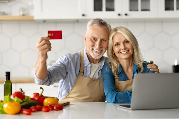 E-Commerce Concept. Senior Couple Holding Credit Card And Using Laptop In Kitchen, Smiling Elderly Spouses Making Online Shopping Or Recommending Website For Ordering Groceries, Closeup Portrait