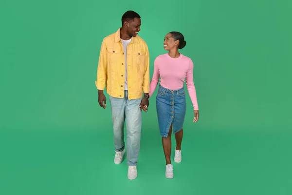 Loving cheerful young african american couple man and woman wearing colorful casual outfits walking over green studio background, holding hands, looking at each other. Love, affection, relationships