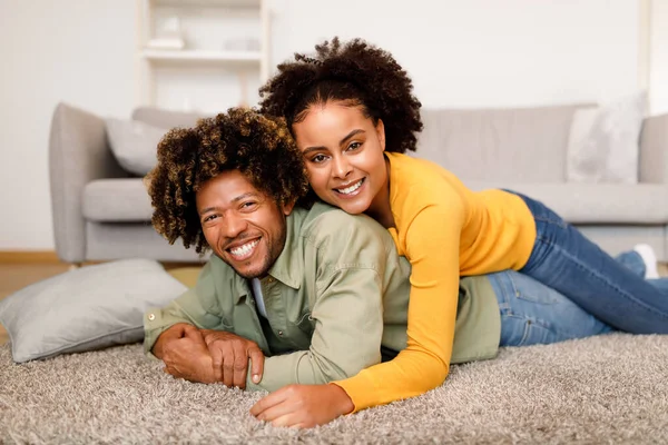 Happy Relationship. Young Black Couple Embracing On Floor And Smiling To Camera At Home, Posing Lying In Modern Living Room Interior. Spouses Enjoying Their Love, Bonding Time On Weekend