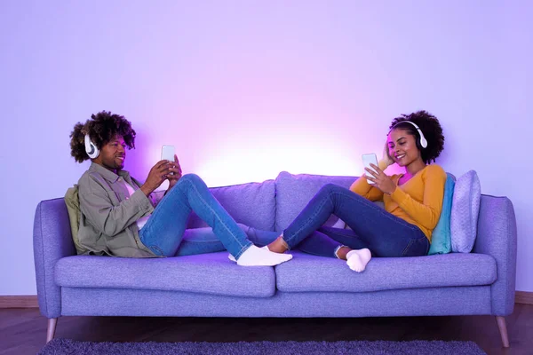 Musical Connection. Happy Black Young Spouses Wearing Headphones, Listening To Music Together on Their Phones, Relaxing on Couch at Living Room Lit With Blue Light on Weekend