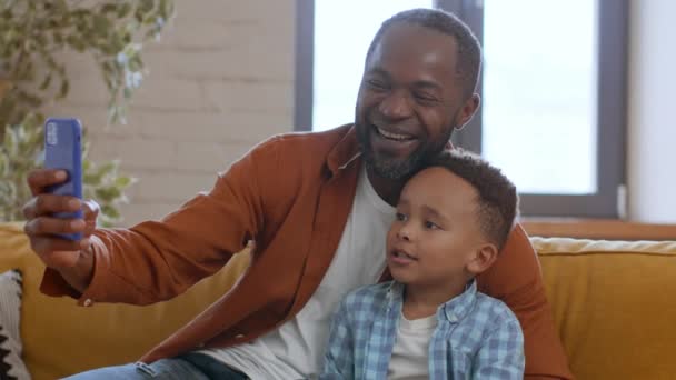 幸せな瞬間をキャプチャする 遊び心のあるアフリカ系アメリカ人の父親と彼の息子は スマートフォンで一緒にセルフィーを作り 自宅でカメラに笑い ショットを追跡 スローモーション — ストック動画