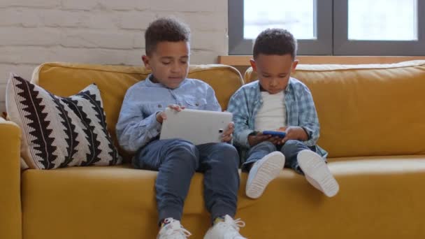 子供とガジェット中毒の問題 2人の小さなアフリカ系アメリカ人の少年がウェブサーフィンし デバイス上でビデオゲームをプレイし 自宅でアクティブなゲームを無視し ショットを追跡し 自由なスペース — ストック動画