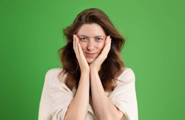 Mutlu Avrupalı genç bayan elleriyle yüzüne dokunuyor, doğal güzelliğin ve mükemmel tenin tadını çıkarıyor, yeşil stüdyo arka planında soyutlanıyor. İnsani duygular, güzellik bakımı, reklam ve teklif, olamaz.