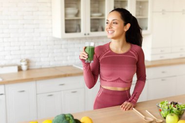 Sağlıklı detoks. Gülümseyen Arap genç kadın yeşil sebzelerden meyve püresi tutuyor, modern mutfakta spor kıyafetleriyle poz veriyor, bardağı tutarken diğer tarafa bakıyor ve sağlıklı içkinin tadını çıkarıyor.