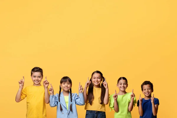 stock image Happy multiracial preteen children cute school aged boys and girls pointing up at copy space for advertisement or text, isolated on yellow background. Great offer for kids