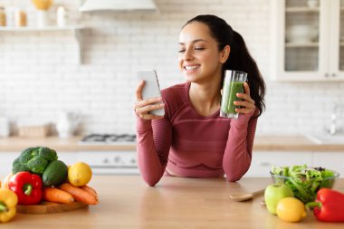 Kilo verme uygulaması. Spor salonundaki kadın akıllı telefon kullanıyor ve evdeki mutfaktaki masada yeşil sebze suyu içiyor. Spor Giysili Kadın Sağlıklı İnce Tarama Tarifleri İnternet 'te
