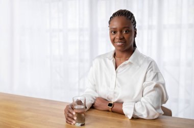 Neşeli, dost canlısı, Afro-Amerikalı, şık giyinen, iş yerinde poz veren, masasında oturan, elinde bir bardak su tutan ve kameraya gülümseyen, reklam için fotokopi çeken güzel kadın.