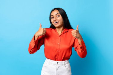 Onaylandı. Ortadoğulu heyecanlı genç bayan, iki eliyle baş parmağını kaldırıyor, harika bir teklif sunmayı seviyor, mavi arka planda poz veriyor, kameraya gülümsüyor. Stüdyo Çekimi