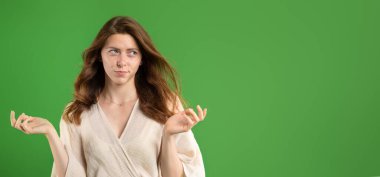 Kafası karışık genç Avrupalı kadın kollarını yana yana açıyor, ne seçeceğini bilmiyor, boş alana bakıyor, yeşil stüdyo arka planında izole edilmiş. İnsani duygular, reklam ve teklif, soru