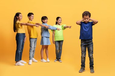 Okul çağında zorbalık. Sarı arka planda izole edilmiş, üzgün çocuğu işaret eden çok ırklı, küçük çocuklar. Zavallı çocuk sınıf arkadaşlarının alaylarından muzdarip, kulaklarını tıkamış.