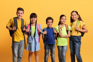 Eğitim, Okul Konsepti. Çeşitli etnik çeşitlilikteki çocuklar kameraya gülümseyen, günlük kıyafetler giyen ve sırt çantası taşıyan, sarı turuncu renkli stüdyo arka planı olan mutlu çocuklar.