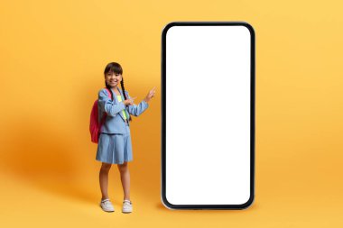 Çocuklar için eğitici mobil uygulama, çevrimiçi öğrenme. Sırt çantalı, beyaz ekranlı, sarı arka planlı, boş ekranlı kocaman bir telefonun yanında duran sevimli, neşeli Asyalı kız.