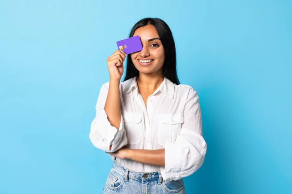 Great Bank Offer. Cheerful Young Indian Lady Holding Bank Credit Card Closing Eye, Smiling To Camera While Advertising Financial Service, Standing Over Blue Studio Background