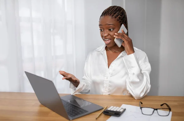Angry young black woman in formal outwear manager sitting at table, looking at laptop screen and gesturing, businesswoman have phone convetsation with assistant, office interior, copy space