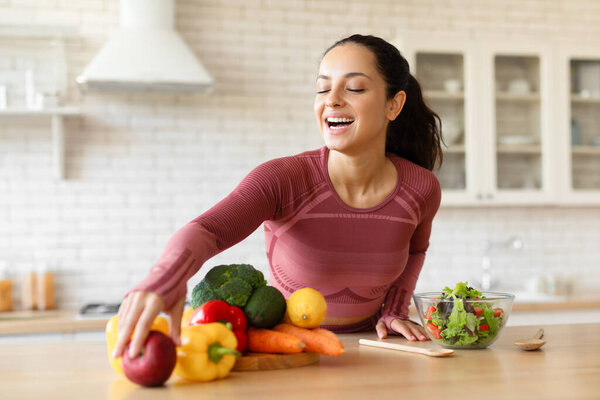 Happy young sporty lady preparing healthy vegetable salad in kitchen interior, enjoys cooking food routine at home, taking fresh fruits and vegatables while preparing dinner. Weight loss nutrition