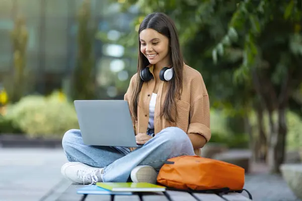 オンライン教育 学生ライフスタイルのコンセプト 陽気で魅力的な若い女性学生は ベンチに座って ラップトップコンピュータ ワイヤレスヘッドフォン コピースペースを使用して 公園で宿題をしている — ストック写真