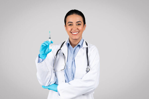 Glad brazilian woman doctor in white coat and gloves showing syringe and smiling, posing isolated on grey studio background. Professional work, medicine and health care, immunization and treatment