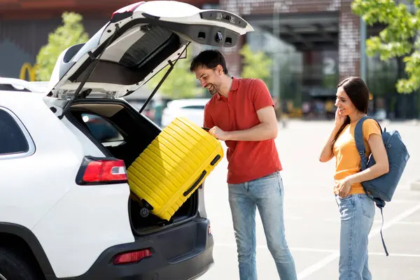 Happy caucasian millennial couple going car trip together. Cheerful young man and woman in casual travellers getting ready before journey by auto, loading vehicle trunk with luggage, stand outdoors