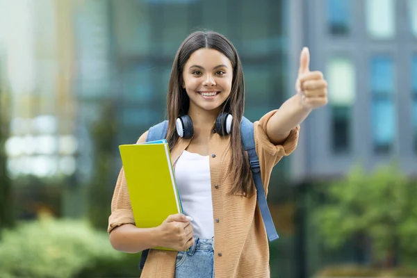 在大学校园里穿着休闲装 背着背包 带着无线耳机 拿着笔记本 露出大拇指的漂亮的年轻黑发女学生 学生生活方式 — 图库照片