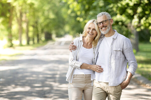 Beautiful mature spouses hugging outdoors while walking in summer park, happy senior couple embracing and looking away, older husband and wife enjoying time together, having romantic date outside