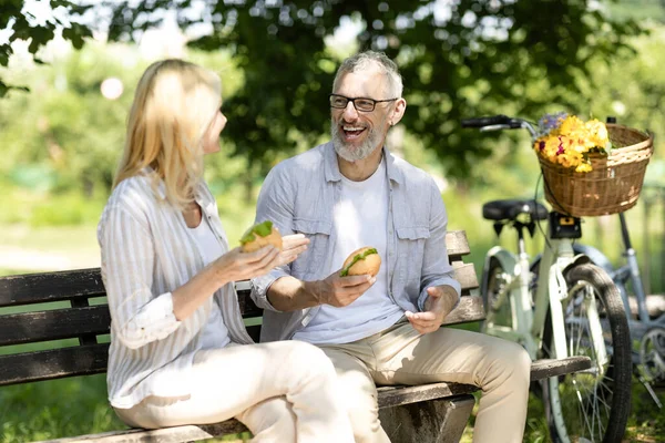 Happy Senior Couple Eating Sandwiches While Sitting On Bench In Park, Romantic Mature Spouses Having Lunch Outdoors And Laughing Together, Cheerful Older Man And Woman Enjoying Picnic Outside