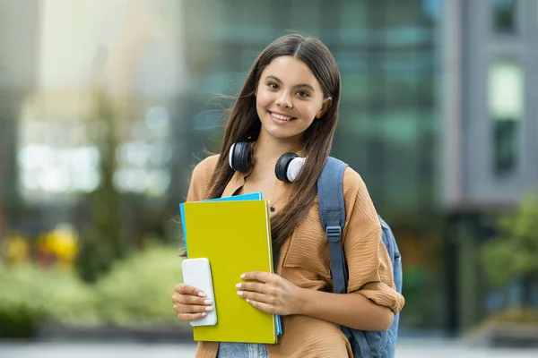 大学や大学のキャンパスでアウトドアでポーズする陽気で美しい若い女性学生 バックパックを運ぶ スマートフォンやコースの本を持って コピースペース ぼやけた背景 教育コンセプト — ストック写真