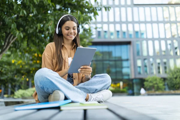 Havalı, uzun saçlı, şık giyinen genç bir bayan parkta oturmuş dijital tablet ve kablosuz kulaklık kullanıyor, online ders izliyor, boş alan izliyor. E-eğitim