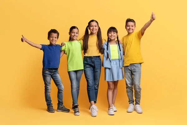 stock image Cool cheerful diverse multiracial preteen kids having fun isolated on yellow background, embracing, showing thumb up and smiling at camera. School aged boys and girls posing together, full length