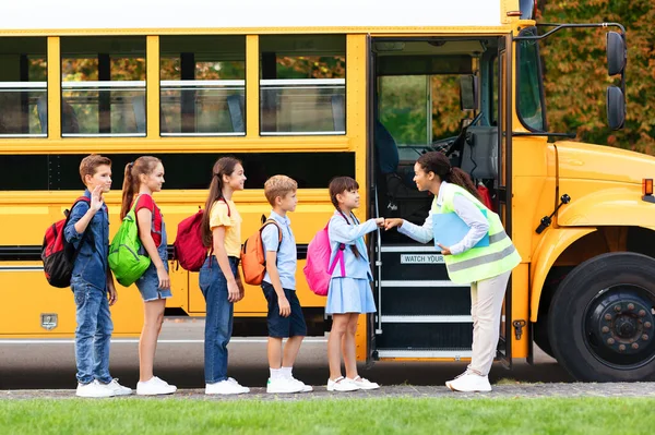 Black female teacher giving fist bumps to students near school bus, smiling assistant lady in uniform interacting with kids while they boarding schoolbus, group of pupils ready for trip together