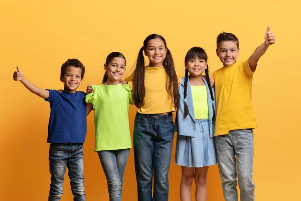 stock image Multiethnic group of school aged kids in casual outfit cheerfully smiling at camera, embracing and showing thumb up posing isolated on yellow background. Happy childhood, kids, friendship concept