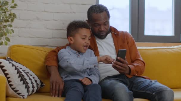 オンラインエンターテイメント かわいいアフリカ系アメリカ人の少年ウェブサーフィン彼の父親とソーシャルメディア スクロールコンテンツと笑い 一緒にソファーに座って ショットを追跡 スローモーション 無料スペース — ストック動画