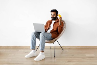 Online Alışveriş Teklifi. Laptop bilgisayarı ve banka kartı ile gülümseyen Orta Doğulu adam internet alışverişi yapıyor, web sitesinde ödeme yapıyor, evdeki beyaz duvarın yanındaki beyaz duvarda oturarak eğleniyor.