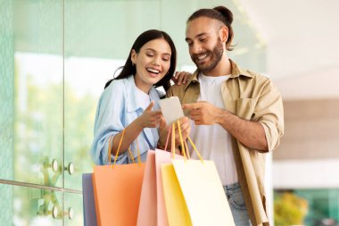 Çantalı neşeli genç Avrupalı çift alışveriş merkezindeki akıllı telefonlara bakarak alışveriş ve boş zamanın tadını çıkarıyor. Satılık uygulama, alışveriş bağımlıları kazanır, şehirde nakit para, indirim ve cihaz olur