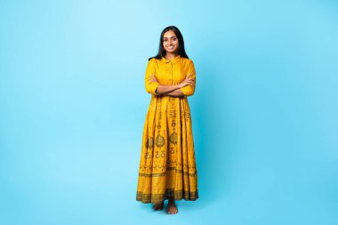 Çekici Hintli genç kadın geleneksel sarı elbiseli, nakışlı, ellerini çapraz yapıp mavi stüdyonun arkasında duran, kameraya gülümseyen. Tam Uzunluk