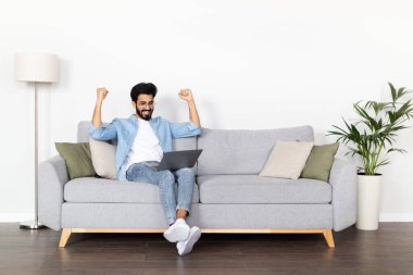 Heyecanlı, mutlu, mutlu, mutlu, bin yıllık Hintli adam koltuğunda oturmuş dizüstü bilgisayar kullanıyor. Doğu 'lu genç adam başarıyı kutluyor, online borsa ve hisse senedi ticareti yapıyor