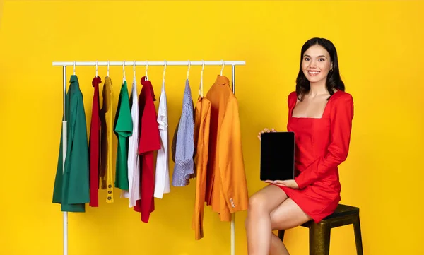 オンラインショッピング 微笑む若い女性は トレンドの服でラックの近くに座っている間 白いデジタルタブレットを表示し ウェブサイトやアプリの広告 モックアップのための黒いデバイス画面をデモンストレーション — ストック写真