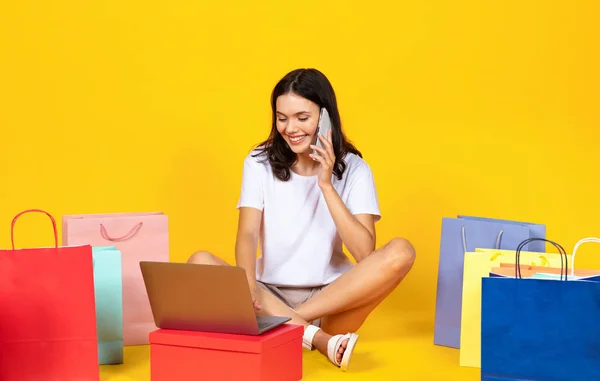 快乐的年轻女人 带着购物袋和笔记本电脑 用手机聊天 微笑着购物的女人 上网购物 确认网上订单 上网购物 坐在黄色工作室的背景上 — 图库照片