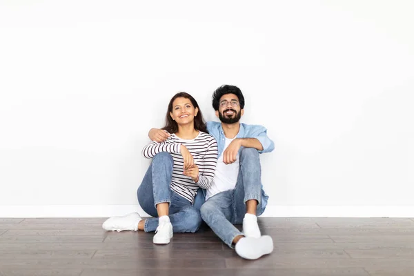 Happy positive beautiful loving young indian couple buying new house, sitting on floor in empty room in apartment, looking at copy space above them on white wall. Mortgage for millennials, rent
