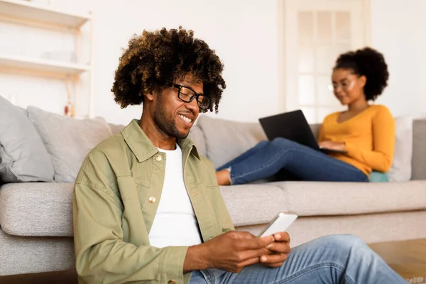 Black Man Surfs Web on Smartphone while His Wife Browses Internet on Laptop Computer, Couple Doing Work Tasks And Enjoying Online Leisure at Home, Wearing Glasses. Family Gadgets. Selective Focus