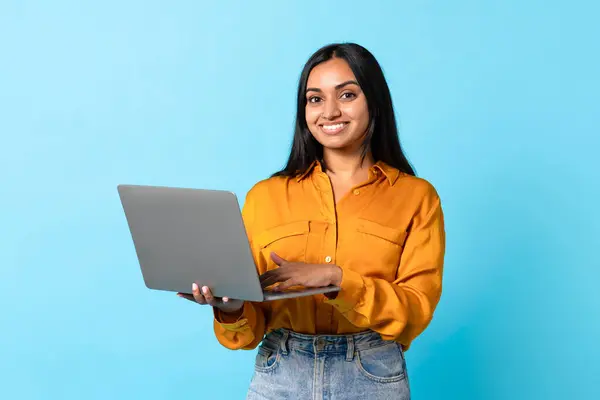 コンピュータソフトウェア広告 ラップトップを使用して陽気なインドの学生の女性 フリーランス作業とEラーニングのためのPcのガジェットとポジショニング ブルースタジオの背景に立って カメラに笑顔 — ストック写真