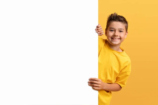 白いプラカードの後ろに隠れている微笑むティーンエイジャーの少年 テキストやデザインのコピースペースを示す幸せな子供の肖像画 水平な空のボード 黄色の背景 モックアップ — ストック写真