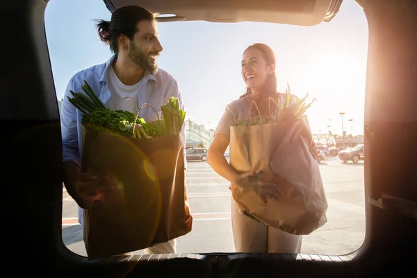 Young latin married couple putting paper bags full of healthy food into car on parking near mall or supermarket. Spouses loading their purchases in car