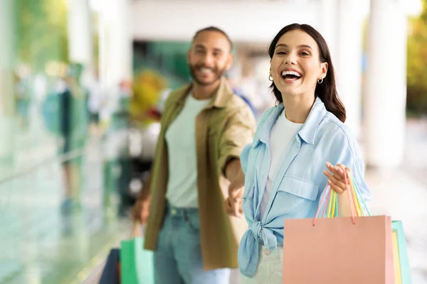 Cheerful young european lady with bags leads man by hand, enjoy shopping, walk and free time in mall. Shopaholics at sale, discount season, lifestyle and purchases, emotions