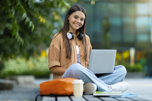 大学毕业后 坐在公园的长椅上 用手提电脑和无线耳机 喝咖啡 复制空间 笑着漂亮的黑发女学生 — 图库照片