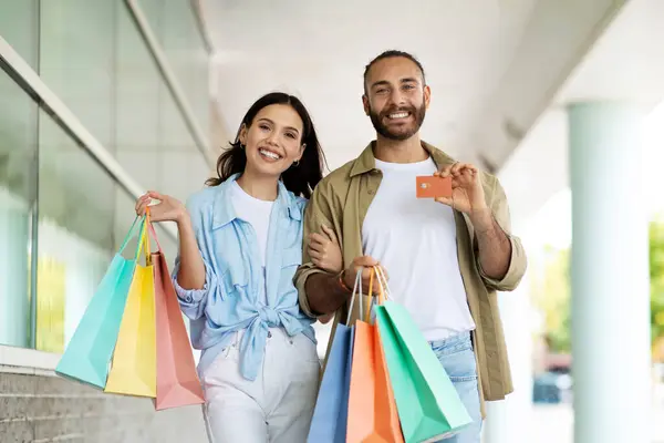 Happy young european man and lady shopaholics with bags show credit card, enjoy shopping and free time in mall. Finance recommendation, money for sale, discount and cashback