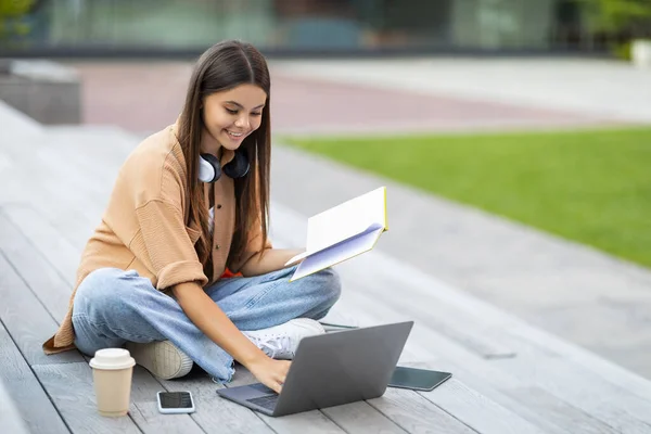 笑顔の魅力的なブルネットスタイリッシュな若い女性の大学生は宿題をしている ベンチに座って 公園でコンピュータを使用して ノートパソコンのキーボードを入力し ノートパッドでメモをチェックし スペースをコピー — ストック写真