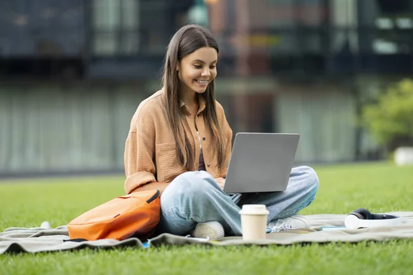 公園でノートブックを使用して涼しく陽気でかわいい若い女性大学生 彼女のラップにラップトップと芝生に座って テイクアウトコーヒーを飲んで 素敵なオンライン教育コースを探して 空のスペース — ストック写真